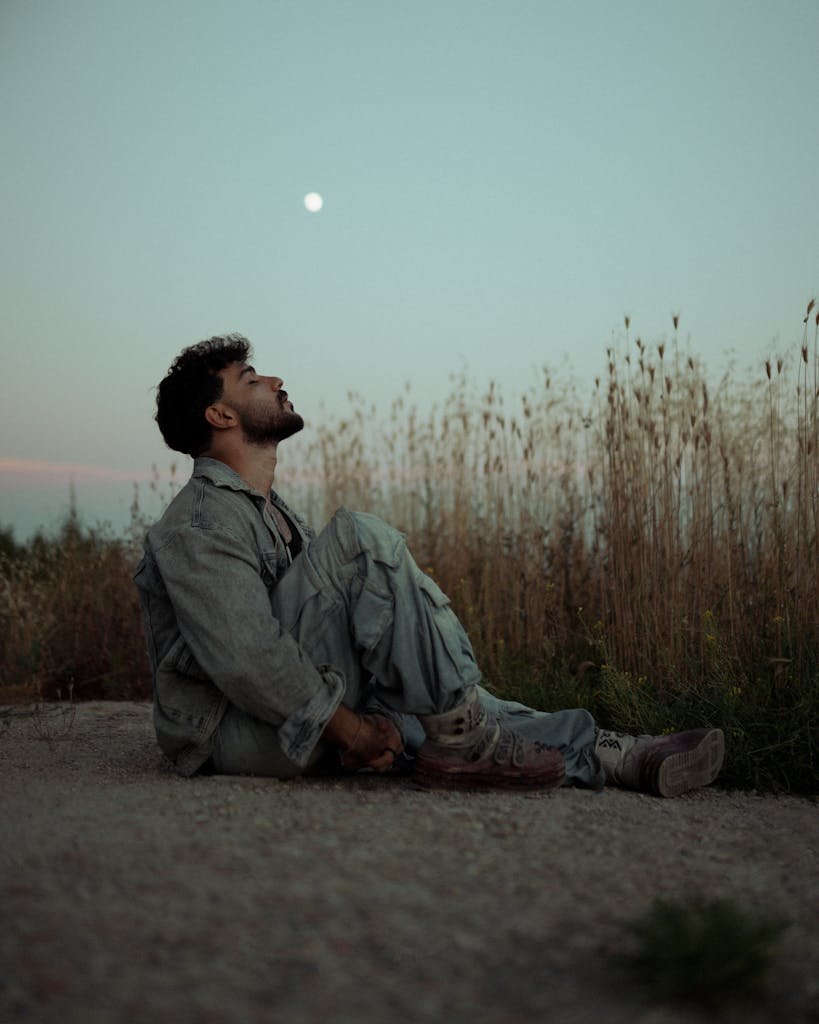 A man in casual attire sits on the ground outdoors, gazing at the moon.