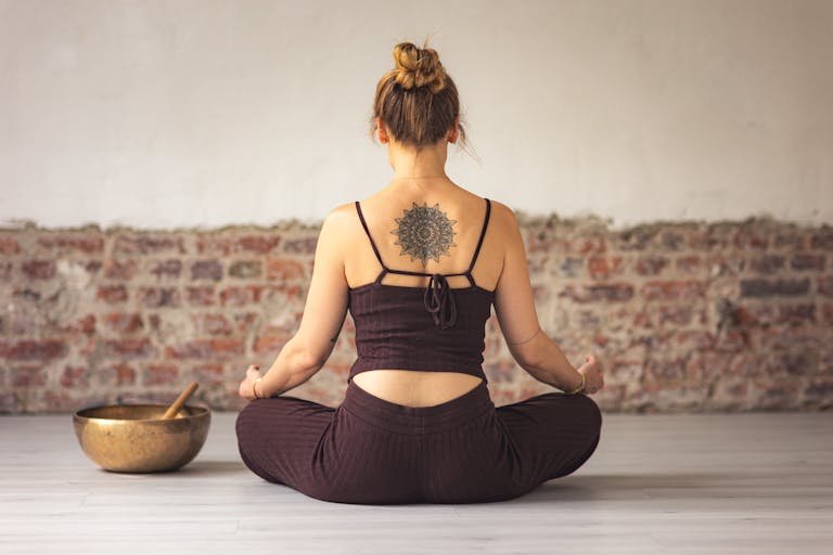 Woman meditating indoors with a tattoo on her back, embracing peace and zen.