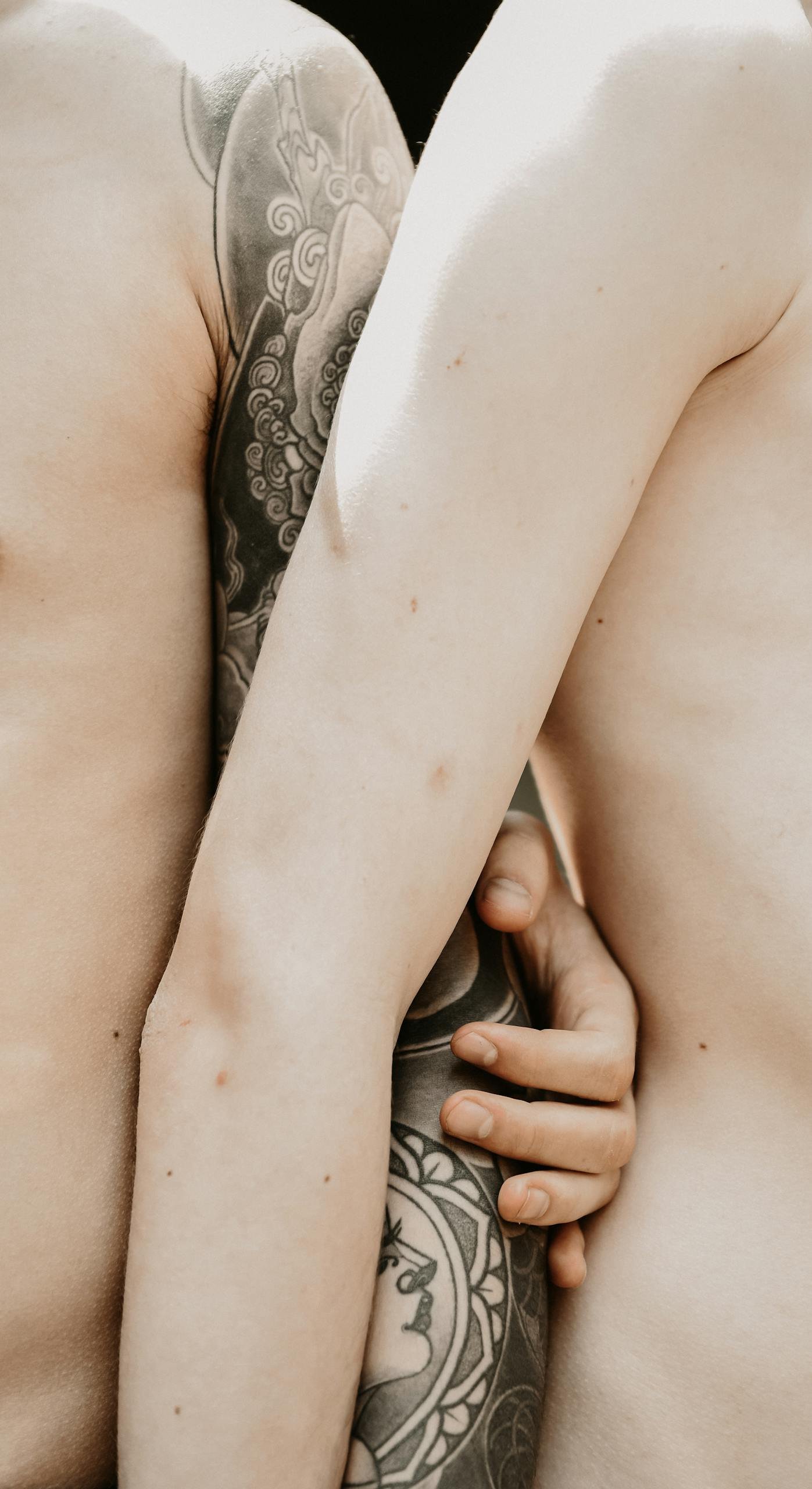 Intimate close-up of two shirtless men embracing, focusing on tattooed arms.