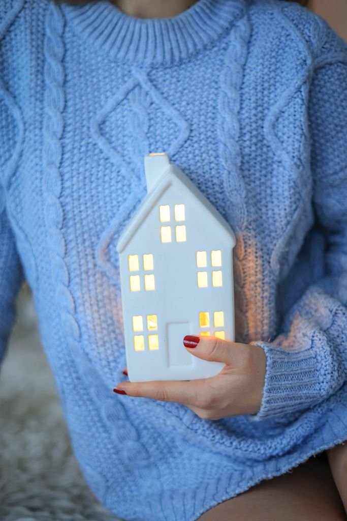 A person wearing a blue sweater holds a glowing miniature ceramic house, symbolizing warmth and comfort.