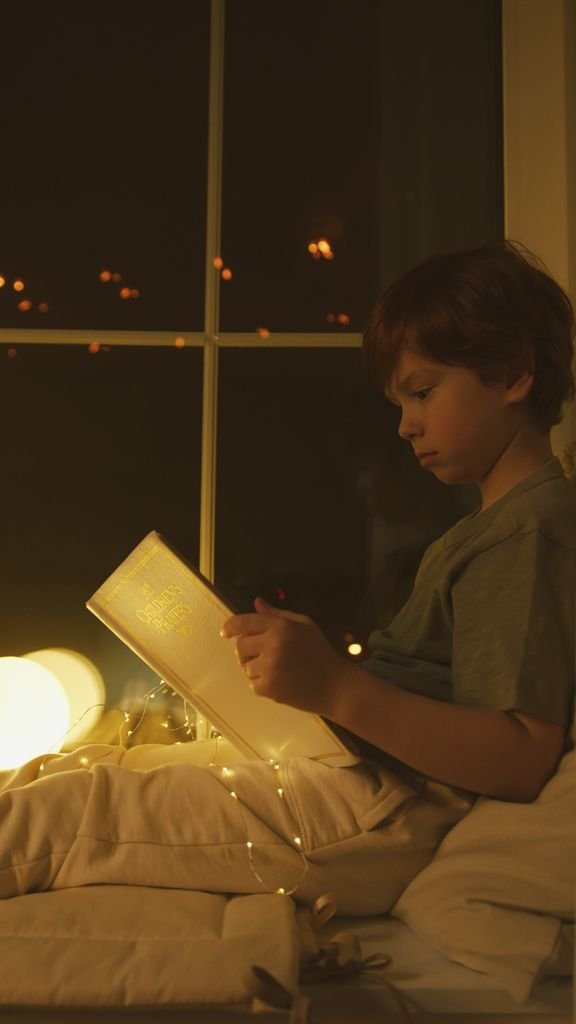 A young boy reads intently by the window, illuminated by warm, cozy lights at night.